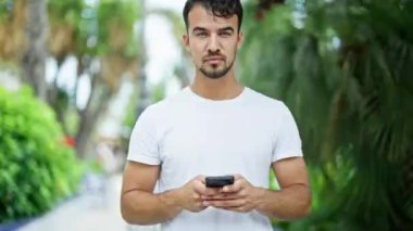 Genç İspanyol adam parkta gülümseyen akıllı telefon kullanıyor.