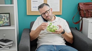 Gri saçlı iş adamı akıllı telefondan konuşuyor ofiste salata yiyor.