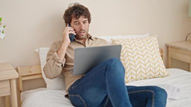 Genç İspanyol adam, yatak odasında dizüstü bilgisayar kullanarak akıllı telefondan konuşuyor.