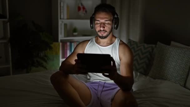 Hombre Árabe Joven Usando Touchpad Sentado Cama Con Auriculares Dormitorio — Vídeo de stock