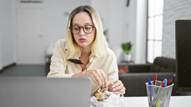 若い金髪女性ビジネスワーカーはオフィスでラップトップとヘッドフォンを使用してサンドイッチを食べる — ストック動画