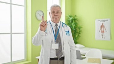 Orta yaşlı, gri saçlı bir doktor ciddi bir ifadeyle klinikte parmağıyla hayır diyor.