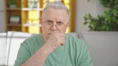 Orta yaşlı, kır saçlı, kanepede oturan, evde ağzı kapalı jest yapan bir adam.