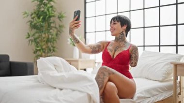 Ampüte kollu, iç çamaşırlı İspanyol kadın yatakta oturmuş, yatak odasında akıllı telefondan selfie çekiyor.