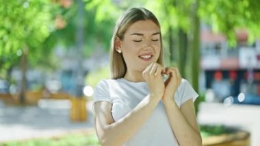 Young blonde woman smiling confident doing heart gesture with hands at park