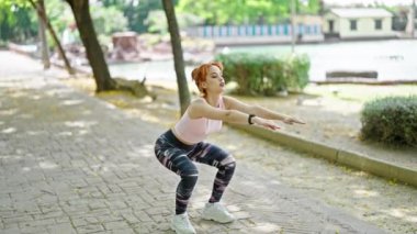 Spor kıyafeti giyen genç kızıl saçlı kadın parkta egzersiz yapıyor.