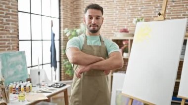 Kollarını kavuşturup sanat stüdyosuna gülümseyen genç Arap sanatçı.
