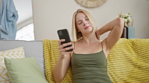 自宅で子宮頸部の痛みに苦しんでいるスマートフォンを使用して若い金髪の女性 — ストック動画