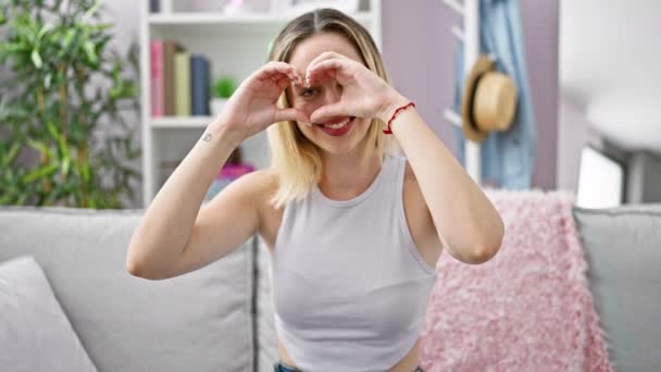 Joven Mujer Rubia Sonriendo Confiada Sentada Sofá Haciendo Gesto Corazón — Vídeo de stock