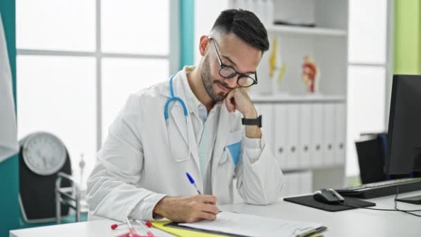 Joven Médico Hispano Tomando Notas Estresado Quitándose Las Gafas Clínica — Vídeo de stock