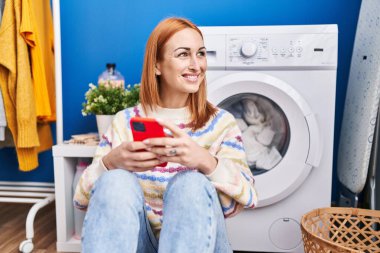 Çamaşır odasında çamaşır makinesi bekleyen beyaz bir kadın akıllı telefon kullanıyor.