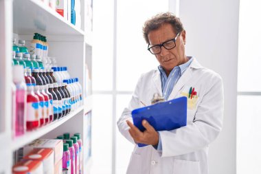 Middle age man pharmacist writing on clipboard looking shelving at pharmacy