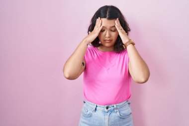 Pembe arka planda duran genç İspanyol kadın, elleri başının üstünde, stresten baş ağrısı çekiyor. migren ağrısı. 