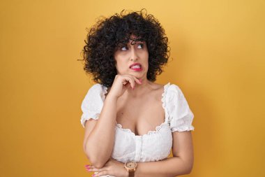 Sarı arka planda duran kıvırcık saçlı genç esmer kadın bir soru hakkında endişeli ve gergin. 