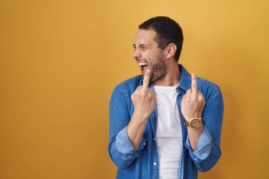 Hispanic man standing over yellow background showing middle finger doing fuck you bad expression, provocation and rude attitude. screaming excited 