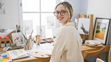 Sanat stüdyosunda gülümseyen genç sarışın kadın sanatçı.