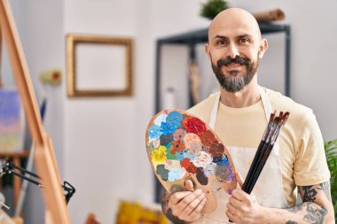 Resim stüdyosunda elinde fırça ve palet tutarak gülümseyen kel bir sanatçı.