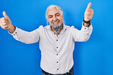 Mavi arka planda gri saçlı orta yaşlı bir adam elleriyle olumlu jest yapmayı onaylıyor, gülümseyerek ve başarı için mutlu. kazanan jest. 