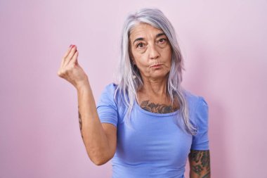 Pembe arka planda dövmeleri olan orta yaşlı bir kadın italyan jestleri yapıyor ve parmaklarında kendinden emin bir ifade var. 