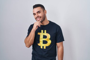 Eli çenesinde Bitcoin tişörtü giyen İspanyol genç adam soru sormayı düşünüyor, düşünceli bir ifade. Gülümseyen ve düşünceli bir yüz. şüphe kavramı. 