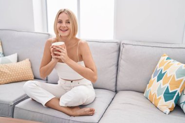 Young blonde woman drinking coffee sitting on sofa at home