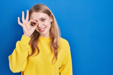 Young caucasian woman standing over blue background doing ok gesture with hand smiling, eye looking through fingers with happy face. 