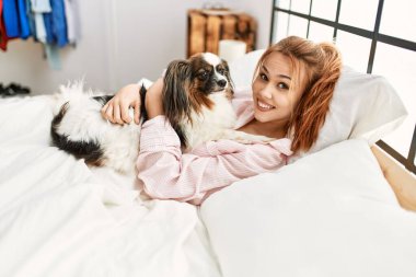 Beyaz bir kadın yatakta uzanıyor. Yatak odasında köpeğe sarılıyor.