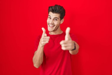 Kırmızı arka planda duran genç İspanyol adam mutlu ve komik yüzlü parmaklarını kameraya doğrultuyor. iyi enerji ve titreşimler. 