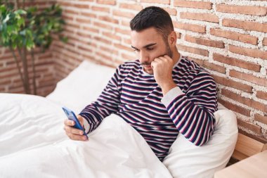 Young hispanic man using smartphone sitting on bed at bedroom