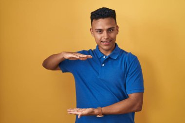 Young hispanic man standing over yellow background gesturing with hands showing big and large size sign, measure symbol. smiling looking at the camera. measuring concept. 