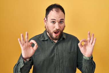 Ayrıca sarı arka planda dikilen sakallı, iri yarı, ispanyol bir adam. Şaşırmış ve şaşırmış görünüyor. Parmaklarıyla onay sembolü yapıyor. çılgın ifade 