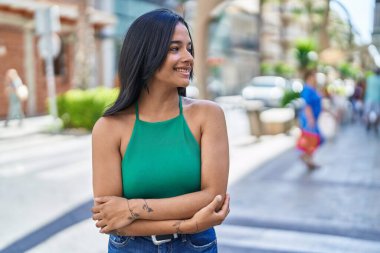 Genç İspanyol kadın gülümsüyor. Kollarını kavuşturup caddede duruyor.