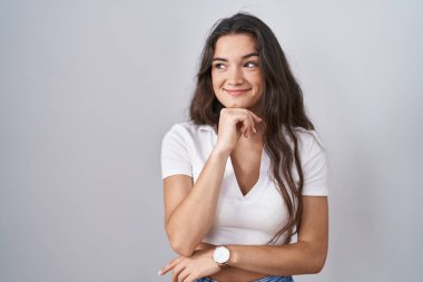 Beyaz arka planda duran genç bir kız, elleri çenesinde soru sorarken, düşünceli bir ifade takınıyor. Gülümseyen ve düşünceli bir yüz. şüphe kavramı. 