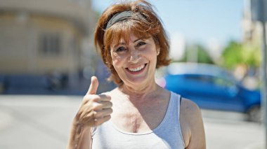Orta yaşlı kadın gülümsüyor. Kendine güveniyor. Sokakta el hareketi yapıyor.