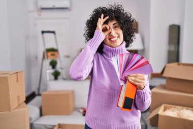 Yeni ev duvarının rengini seçen kıvırcık saçlı, genç esmer kadın gülümsüyor. 