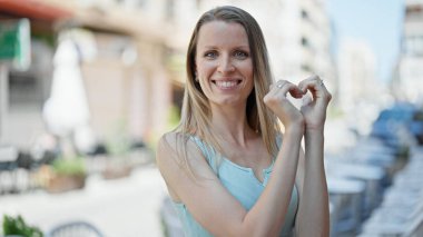 Young blonde woman smiling confident doing heart gesture with hands at street