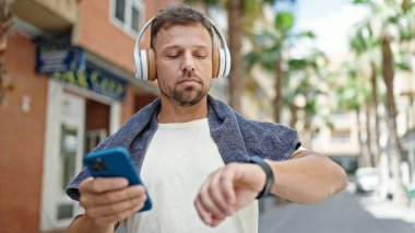 Spor kıyafetleri giyen, müzik dinleyen, akıllı telefon kullanan ve caddede takılan genç bir adam.