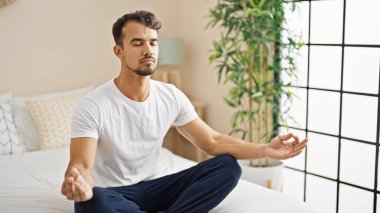Young hispanic man doing yoga exercise sitting on bed at bedroom
