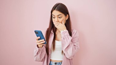 Genç, güzel, İspanyol bir kadın, pembe arka planda sürpriz bir ifadeyle akıllı telefon kullanıyor.