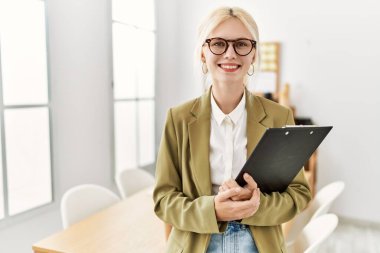 Young blonde woman business worker smiling confident holding clipboard at office
