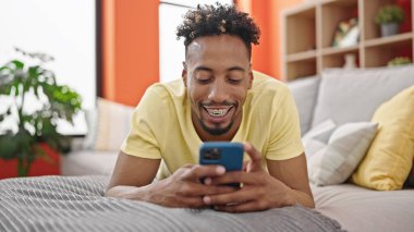 Afro-Amerikalı adam akıllı telefon kullanıyor. Evdeki koltukta yatıyor.