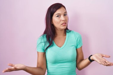 Pembe arka planda duran Latin kökenli genç bir kadın. Kolları açık ve kafası karışık, hiçbir fikri yok.. 