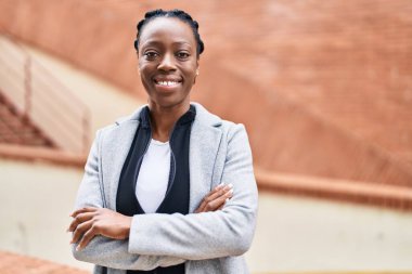 Afro-Amerikalı kadın gülümsüyor. Kollarını kavuşturup caddede duruyor.