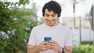 Parkta akıllı telefon kullanan genç latin adam kulaklık takıyor.