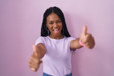 Saçları pembe arka planda dikilmiş afro-amerikan bir kadın elleriyle olumlu jest yapıyor, gülümsüyor ve başarı için mutlu. kazanan jest. 