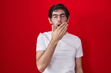 Young hispanic man standing over red background bored yawning tired covering mouth with hand. restless and sleepiness. 