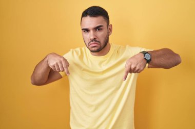 Young hispanic man standing over yellow background pointing down looking sad and upset, indicating direction with fingers, unhappy and depressed. 