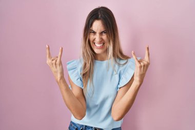 Genç İspanyol kadın pembe arka planda duruyor çılgın bir ifadeyle bağırıyor elleri havada rock sembolü yapıyor. Müzik yıldızı. ağır kavram. 