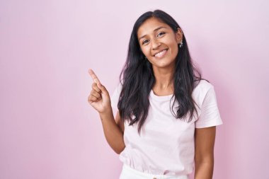 Young hispanic woman standing over pink background cheerful with a smile on face pointing with hand and finger up to the side with happy and natural expression 