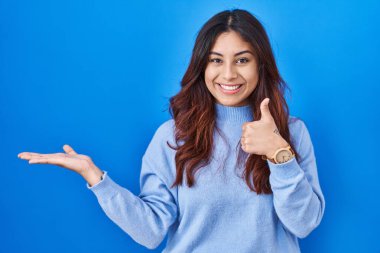 Mavi arka planda duran İspanyol genç kadın avuç içini gösteriyor ve baş parmağıyla iyi bir hareket yapıyor, mutlu ve neşeli gülümsüyor. 